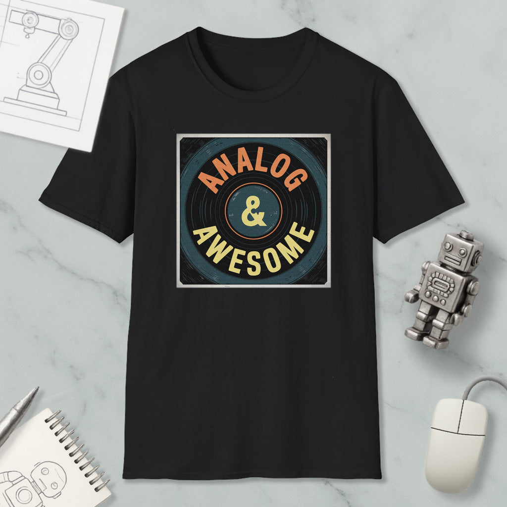 a black t-shirt with a graphic design that says "ANALOG & AWESOME" in a retro style. The t-shirt is placed on a marble surface, surrounded by various objects such as a computer mouse, a robot, and a notebook.