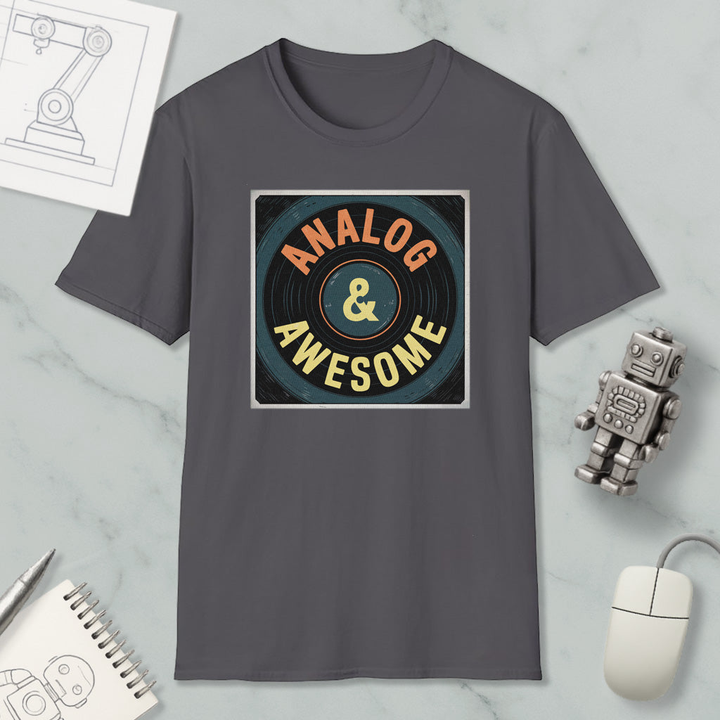 a gray t-shirt with a graphic design that says "ANALOG & AWESOME" in bold letters, surrounded by a circular pattern. The t-shirt is placed on a marble surface, with various objects such as a computer mouse, a robot, and a notebook nearby.