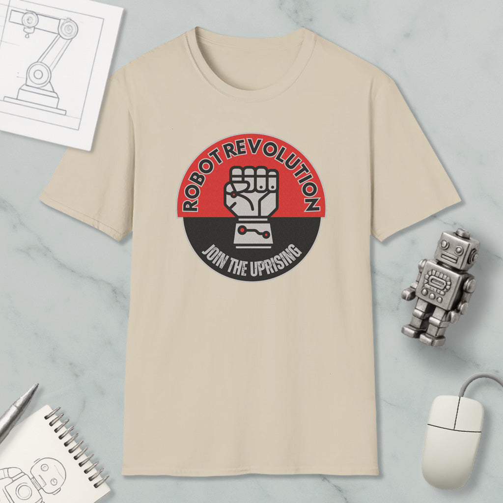 a beige t-shirt with a graphic design that says "Robot Revolution" and "Join the Uprising", along with a fist symbol. The t-shirt is placed on a marble surface, and there are various objects around it, including a computer mouse, a pen, a notebook, and a small robot figurine.