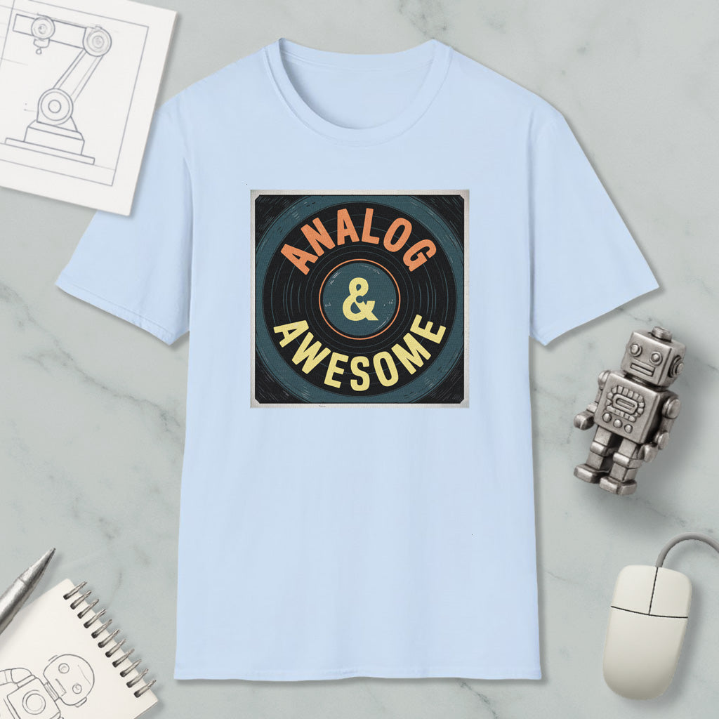 a light blue t-shirt with a graphic design that says "ANALOG & AWESOME" in bold letters. The t-shirt is placed on a marble surface, surrounded by various objects such as a computer mouse, a robot, and a notebook.
