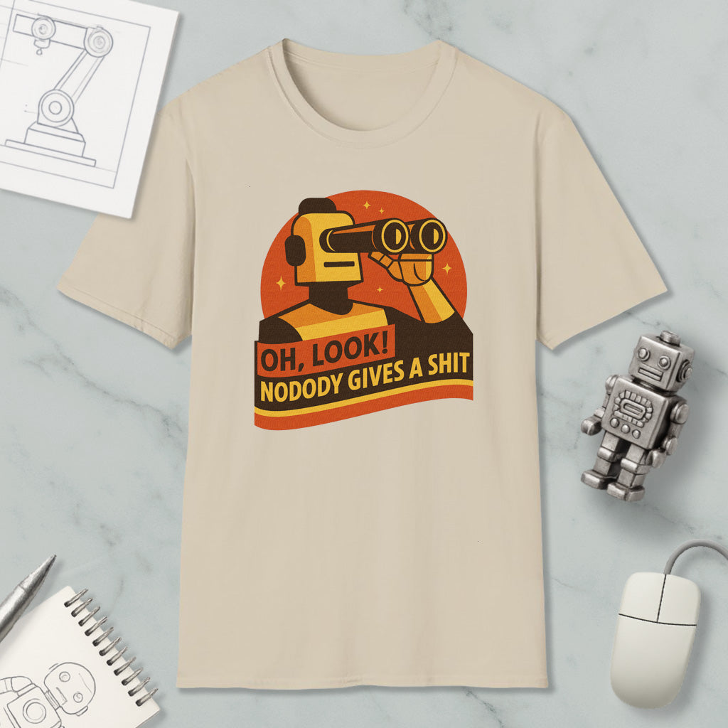 a beige t-shirt with a graphic design depicting a robot character looking through binoculars and holding a sign that says "OH, LOOK! NODODY GIVES A SHIT". The t-shirt is displayed on a marble surface, along with a computer mouse, a pen, and a notebook.