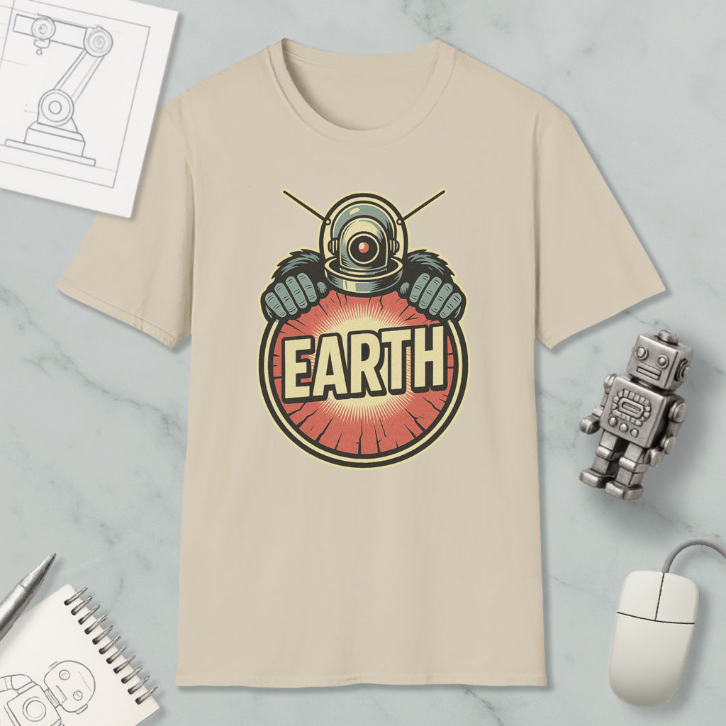 a beige t-shirt with a graphic design of a robot or alien character wearing a helmet and holding a red circle with the word "EARTH" written on it. The t-shirt is placed on a marble surface, along with a computer mouse, a notebook, and a small robot figurine.