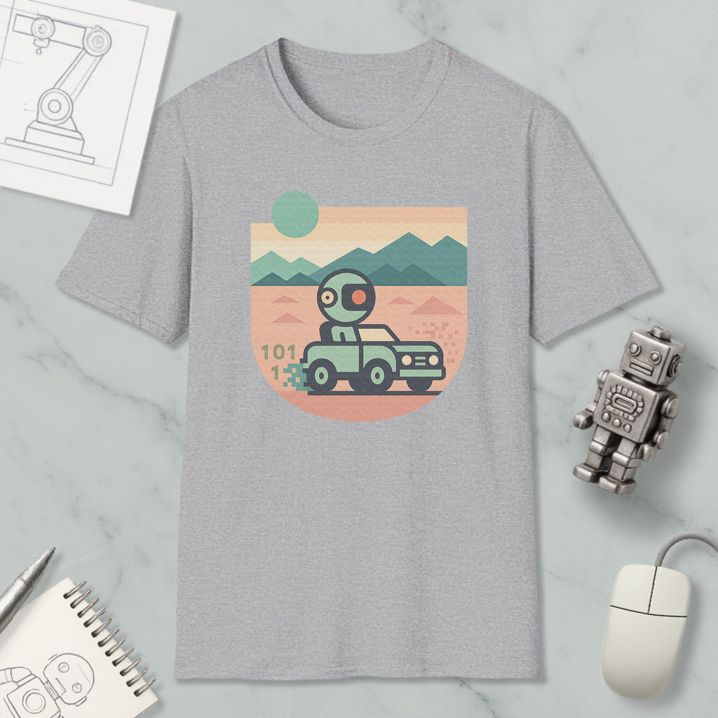 a gray t-shirt with a graphic design depicting a robot driving a car on a desert road, with mountains in the background. The t-shirt is placed on a marble surface, along with a computer mouse, a notepad, and a small robot figurine.