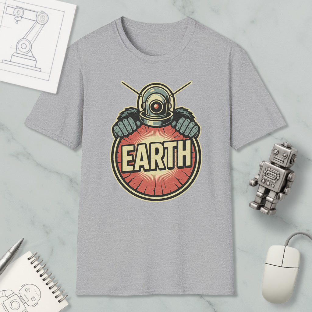 a gray t-shirt with a graphic design of a robot or alien figure holding a red circle with the word "EARTH" written on it. The t-shirt is placed on a marble surface, surrounded by various objects such as a computer mouse, a notebook, and a pen.