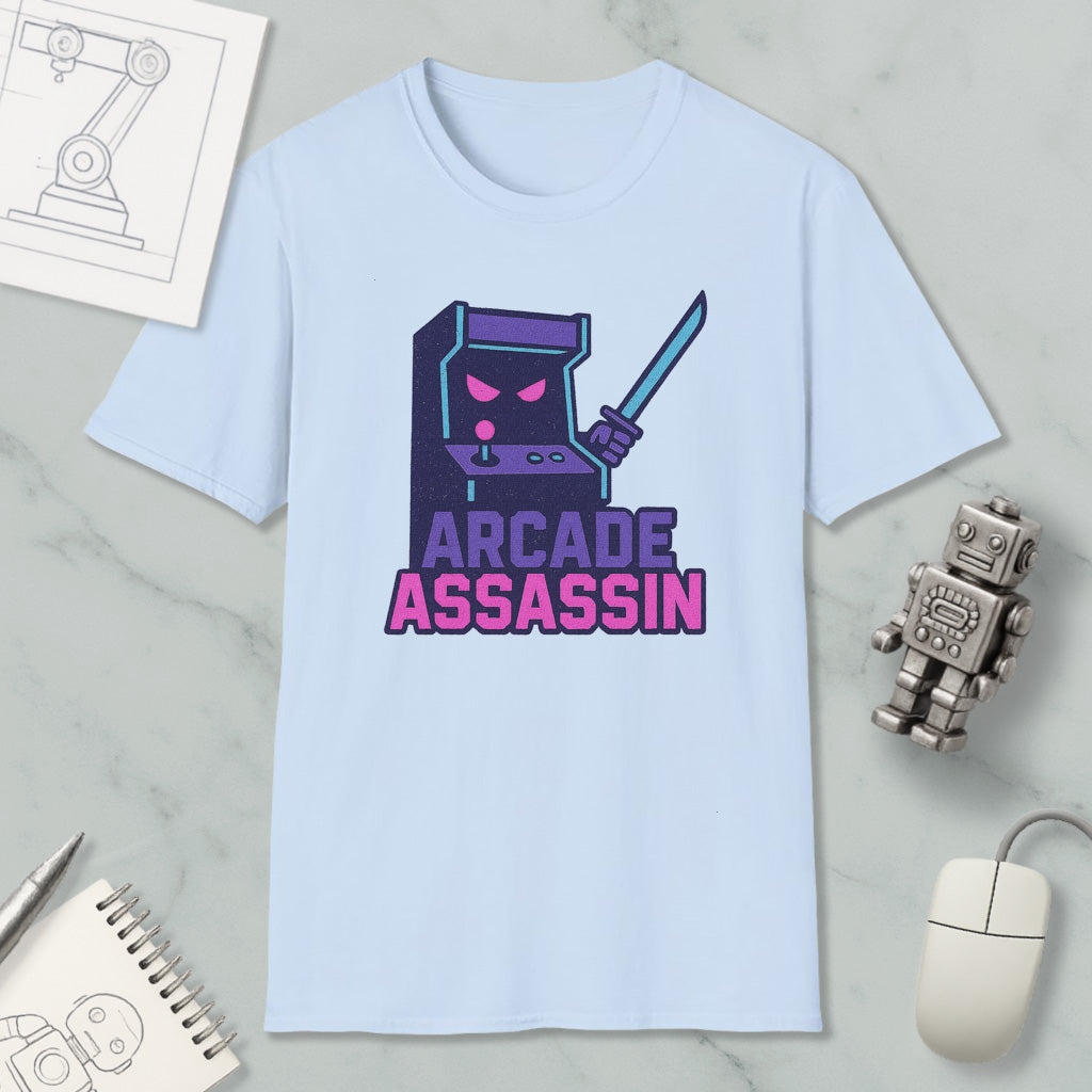 a light blue t-shirt with a graphic design depicting a arcade cabinet character holding a sword and the text "ARCADE ASSASSIN" printed on it. The t-shirt is placed on a marble surface, surrounded by various objects such as a computer mouse, a notebook, and a small robot.