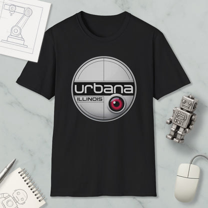 a black t-shirt with a circular logo, a red eye, and the text "urbana illinois" printed on it. The t-shirt is placed on a marble surface, surrounded by various objects such as a computer mouse, a notebook, and a small robot.