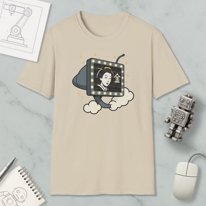 a beige t-shirt with a graphic design depicting a woman's face on a television screen, surrounded by clouds and stars. The t-shirt is placed on a marble surface, along with a computer mouse, a notebook, and a small robot figurine.