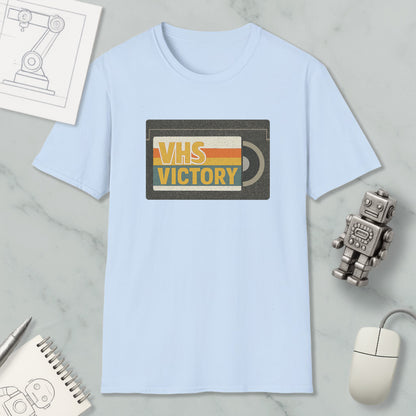 a light blue t-shirt with a graphic design that says "VHS VICTORY" on it, along with a small robot toy and a computer mouse.