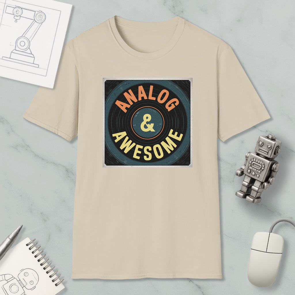 a beige t-shirt with a graphic design that says "ANALOG & AWESOME" in a circular pattern. The t-shirt is placed on a marble surface, surrounded by various objects such as a computer mouse, a robot, and a notebook.