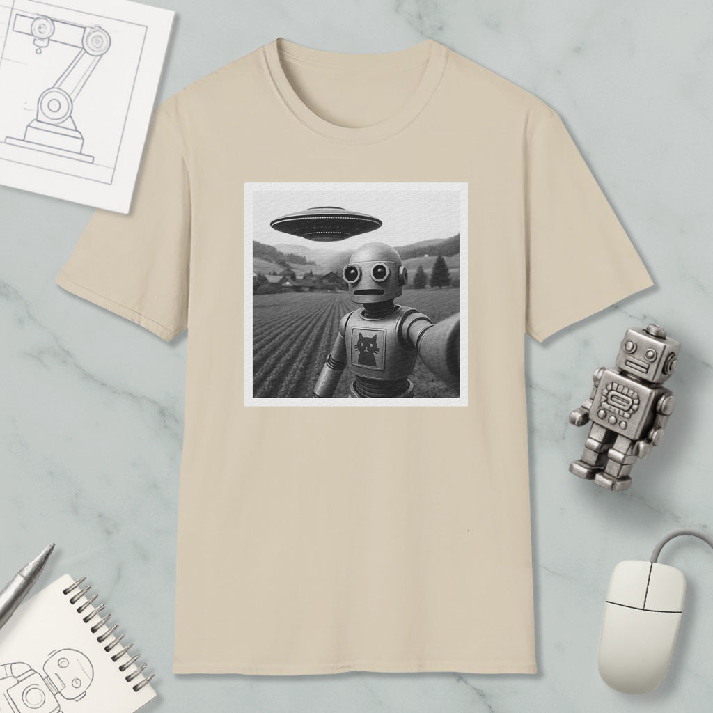 a beige t-shirt with a black and white photograph of a robot standing in a field, with a large spaceship flying above it. The t-shirt is displayed on a marble surface, along with a small robot figurine, a notebook, and a computer mouse.