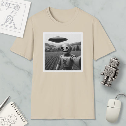 a beige t-shirt with a black and white photograph of a robot standing in a field, with a large spaceship flying above it. The t-shirt is displayed on a marble surface, along with a small robot figurine, a notebook, and a computer mouse.