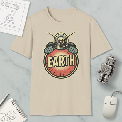 a beige t-shirt with a graphic design of a robot or alien character wearing a helmet and holding a red circle with the word "EARTH" written on it. The t-shirt is placed on a marble surface, along with a computer mouse, a notebook, and a small robot figurine.